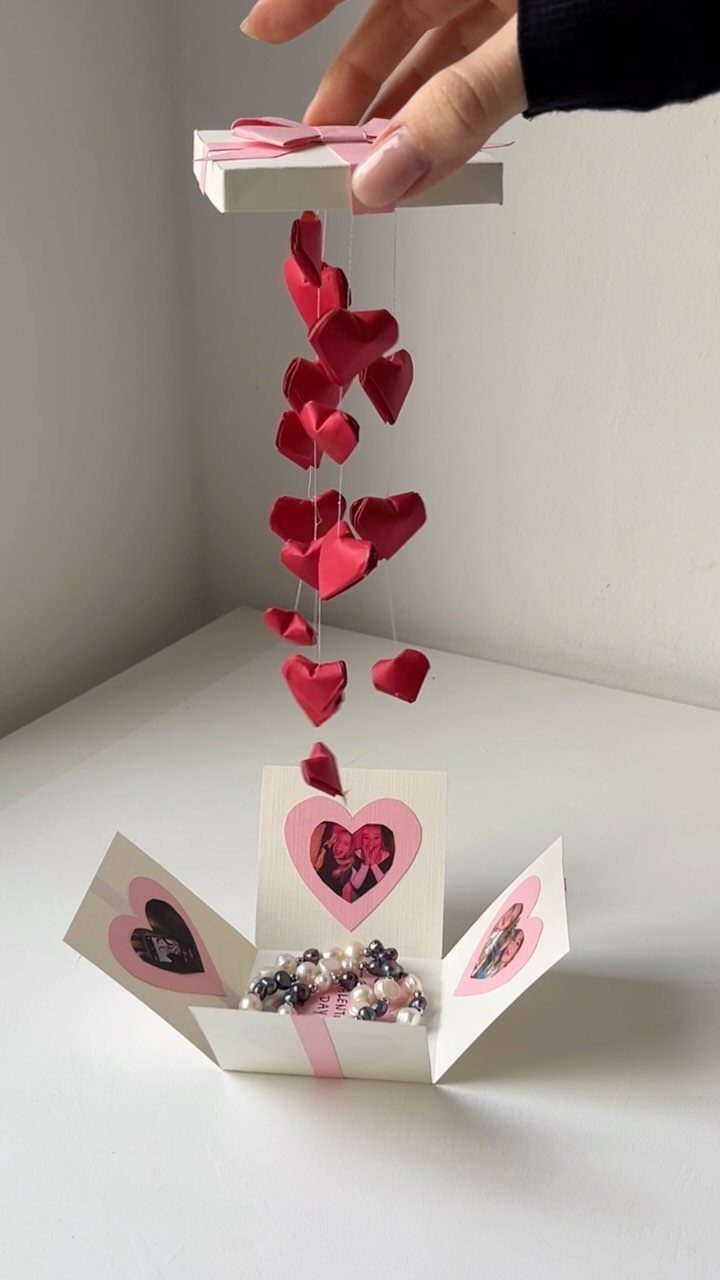 Hand lifting a gift box lid to reveal hanging red hearts and pearls.