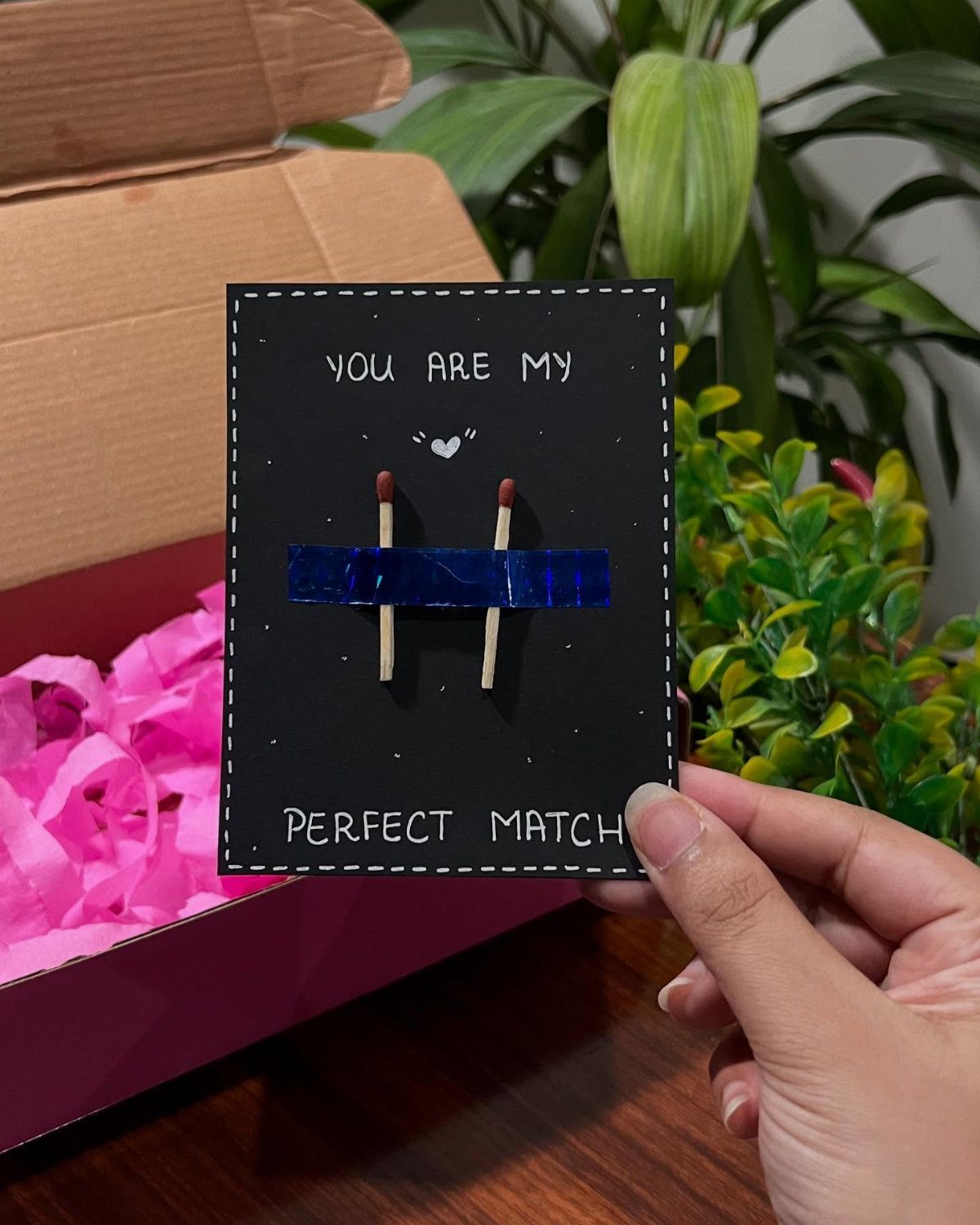 Handmade anniversary card with matchsticks and text held over a pink gift box.