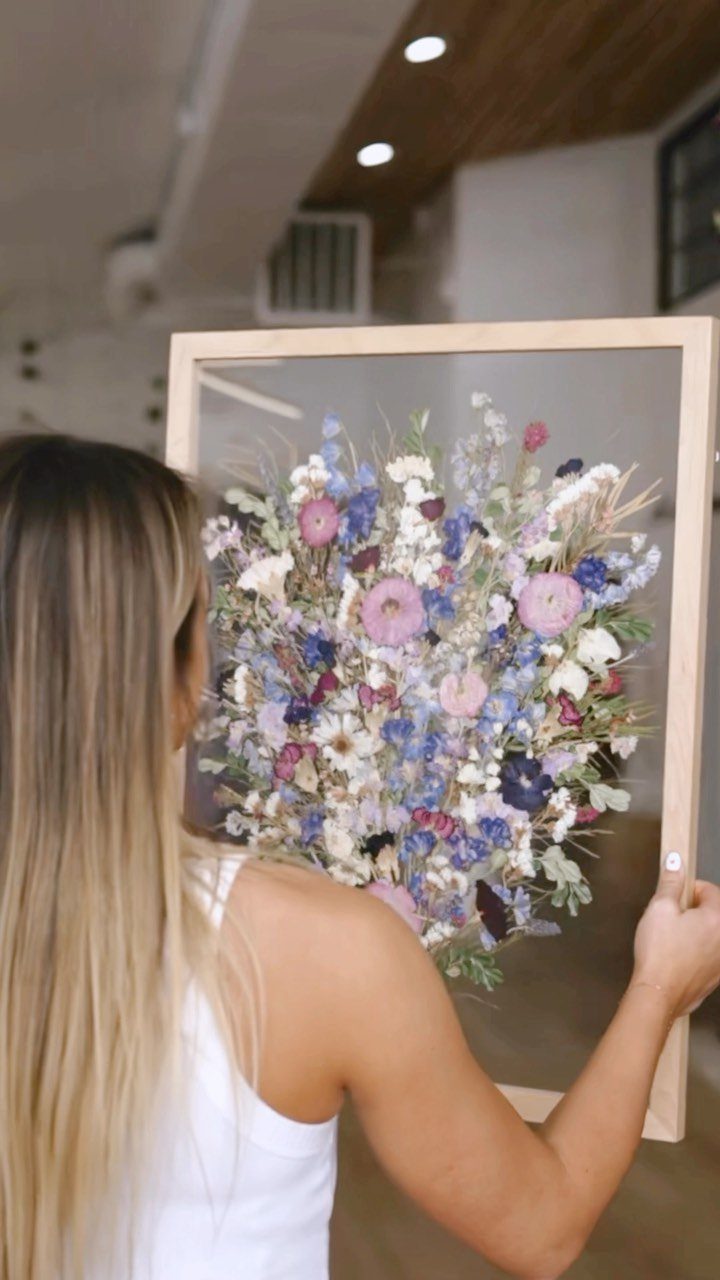 Woman holding a wooden frame with pressed purple and blue flowers inside.