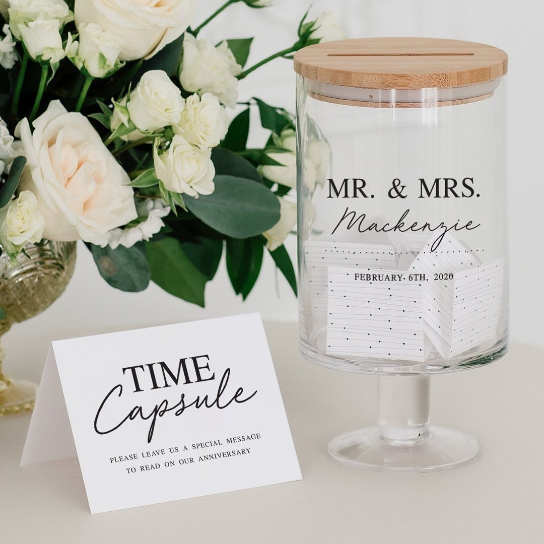 Glass time capsule jar with wooden lid and white slips of paper beside roses