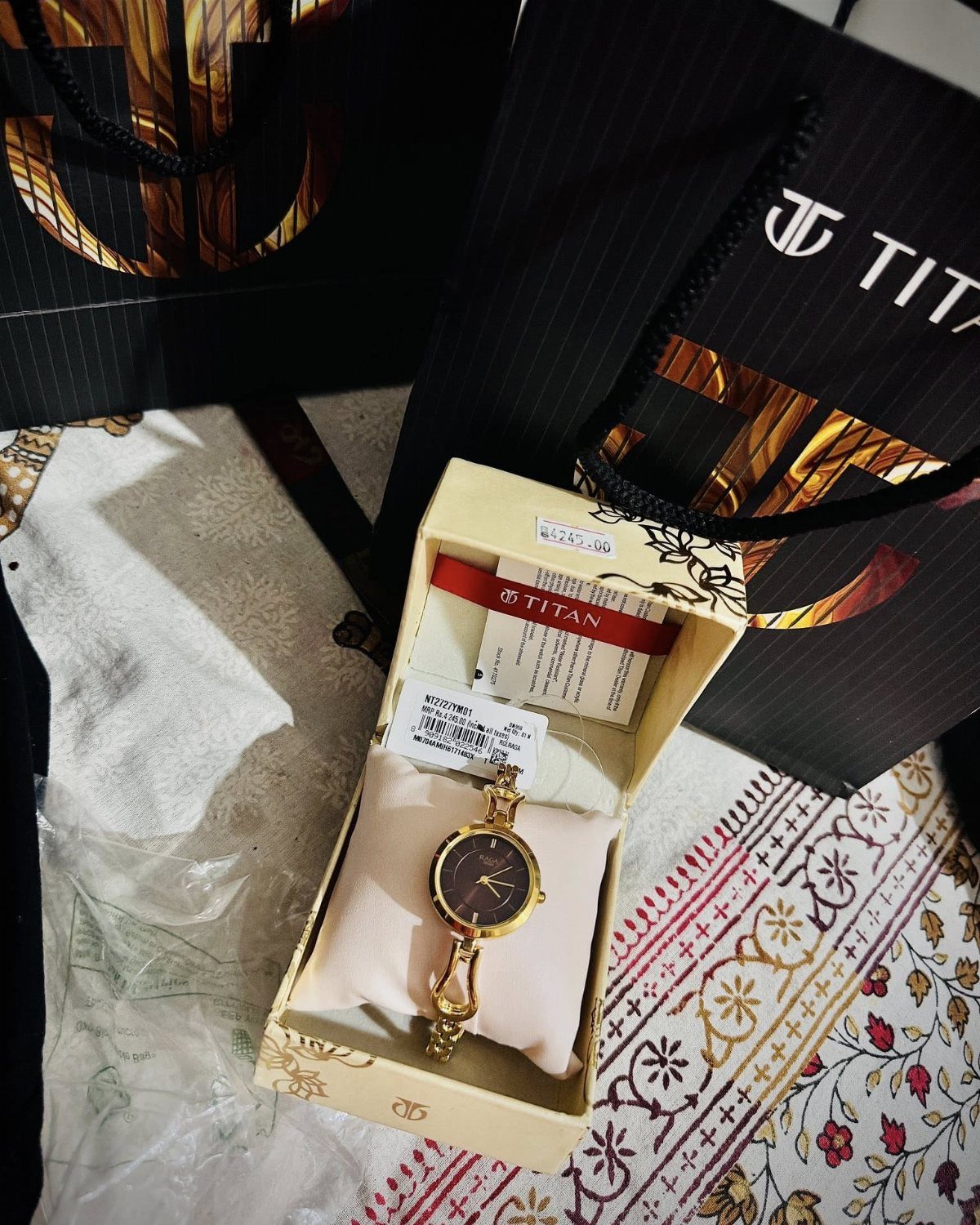 Gold wristwatch in a cream gift box next to black Titan shopping bags.