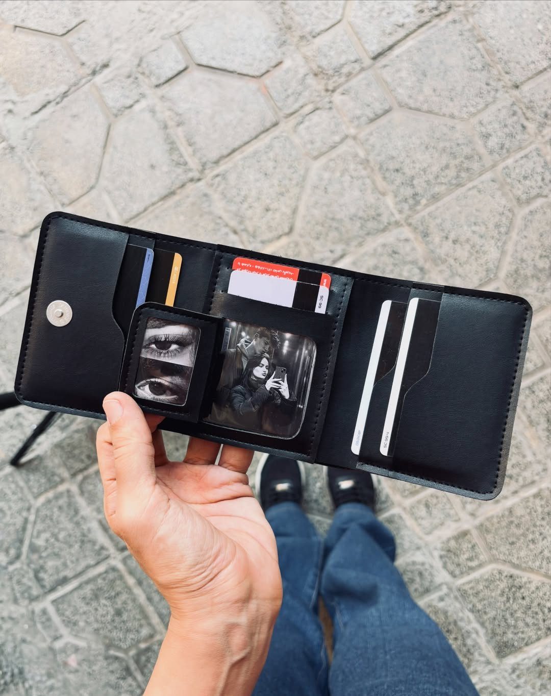 Black leather tri-fold wallet held open showing cards and photos over cobblestone ground