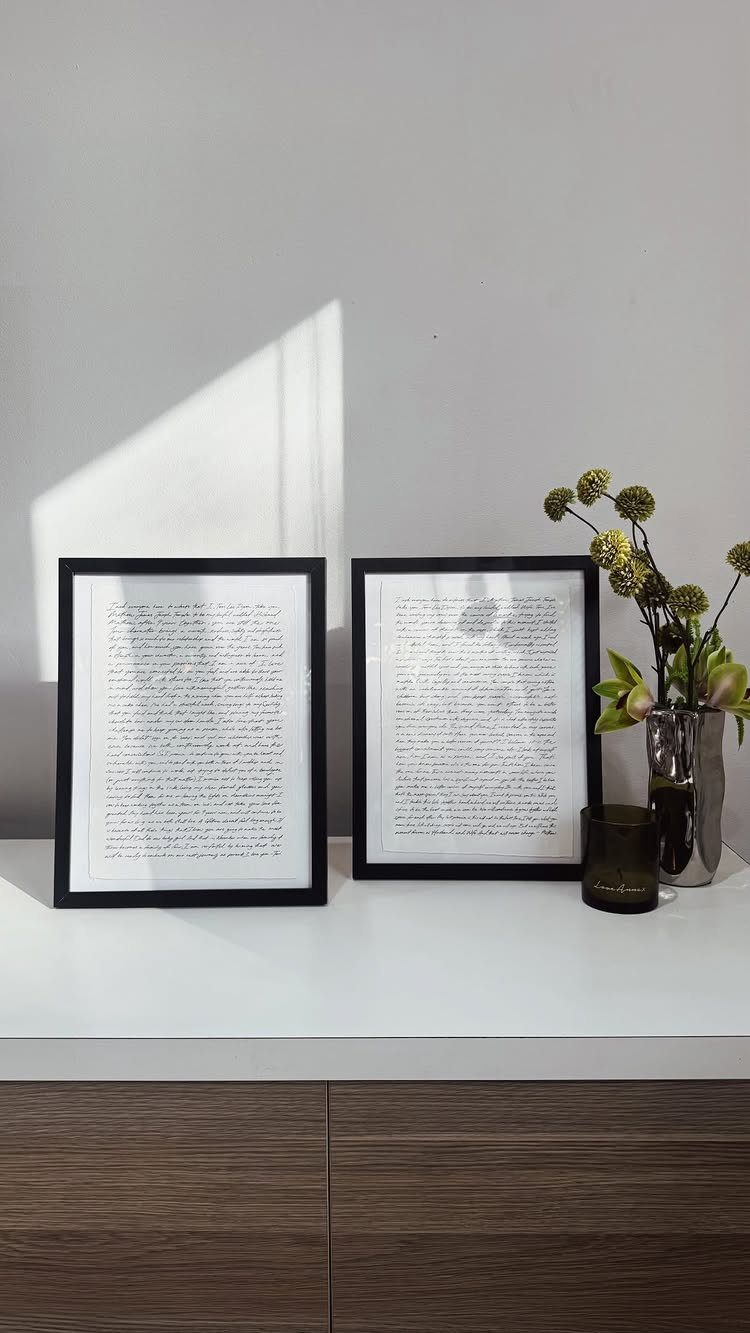 Two black-framed prints of handwritten vows resting on a white table with flowers.