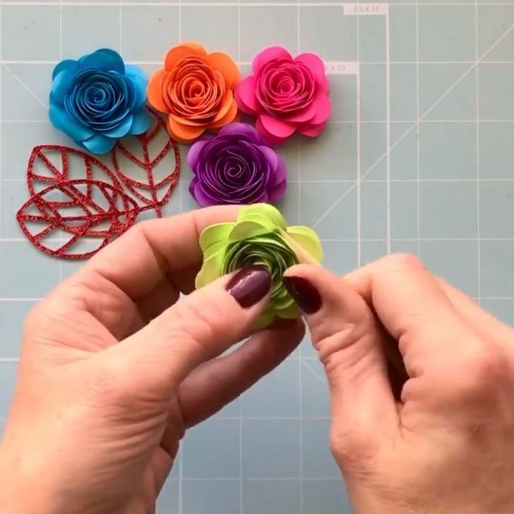 Hands assembling a green paper rose with other colorful paper flowers nearby.