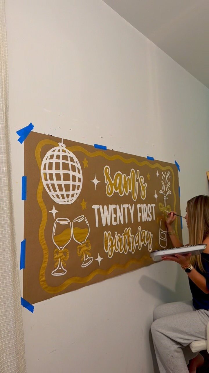 Woman painting a brown 21st birthday banner with a disco ball and champagne glasses.