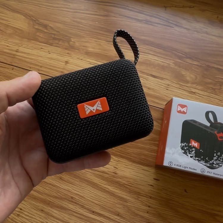 Small black portable Bluetooth speaker with orange logo held over wooden table