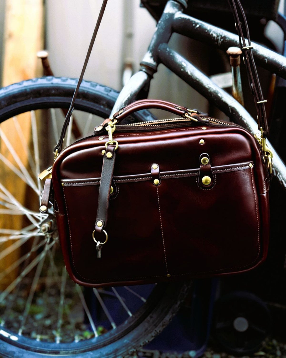 Burgundy leather messenger bag with gold hardware hanging from a black bicycle frame.