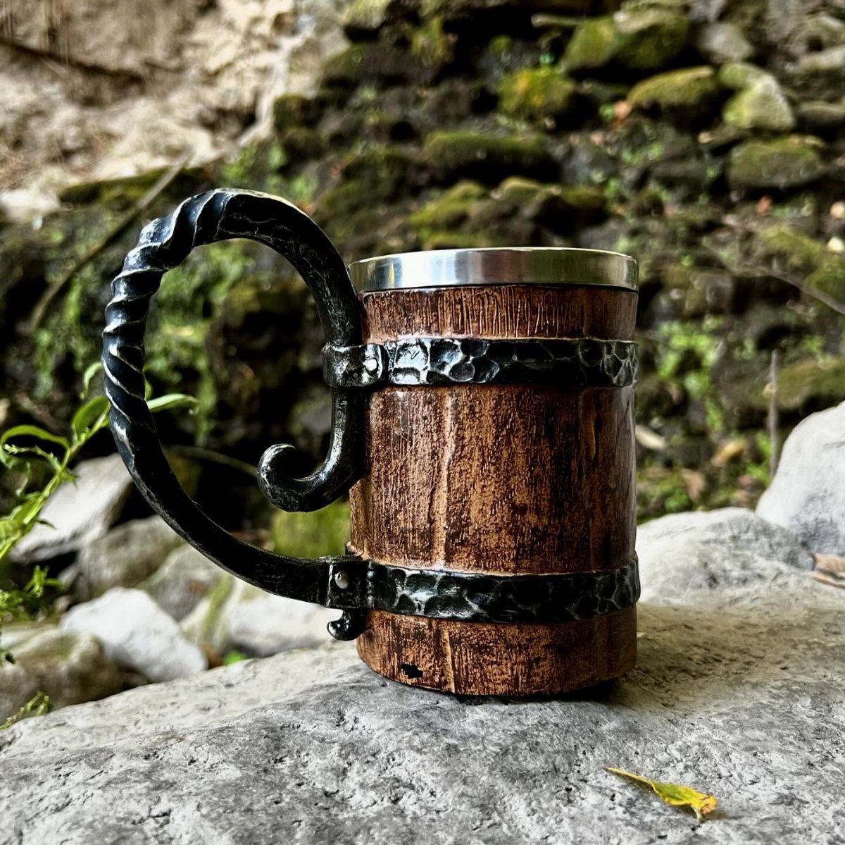 Wooden Viking tankard mug with twisted metal handle resting on a grey rock.