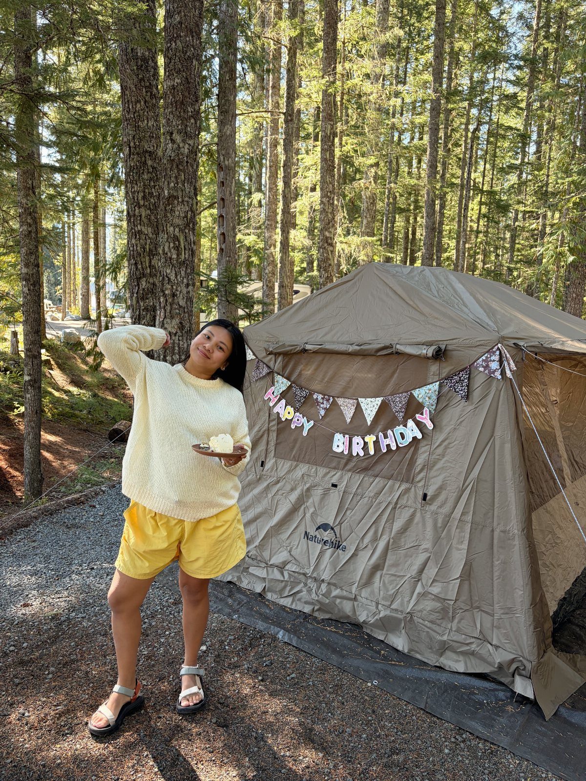 Woman holding a cake next to a birthday decorated tent in a pine forest