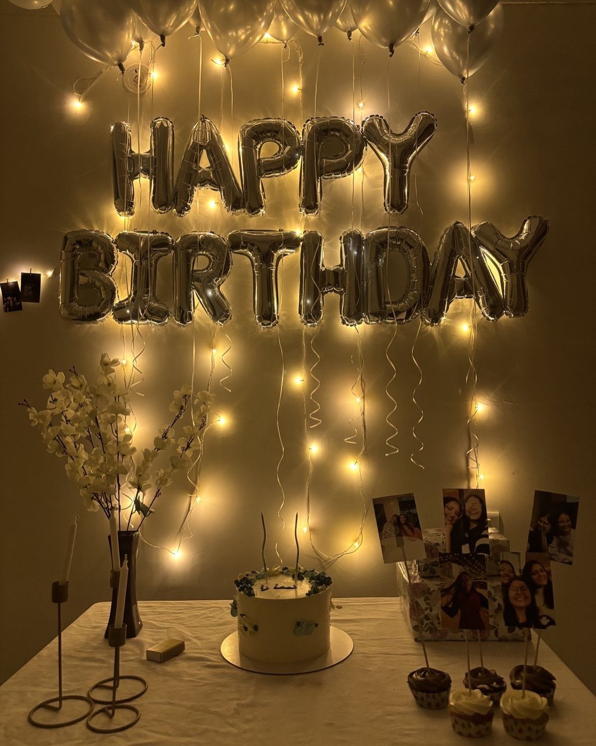 Birthday celebration setup with silver balloons, string lights, cake, and personal photos.