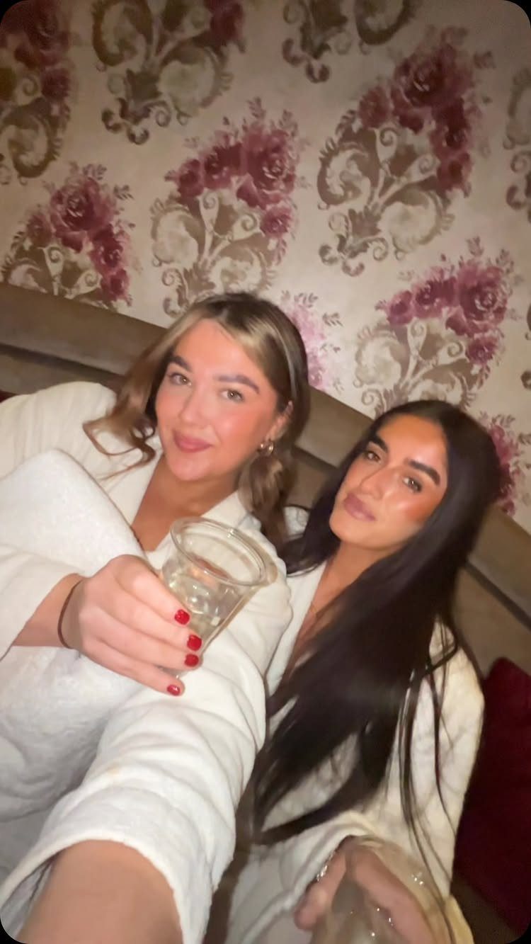 Two young women in white bathrobes smiling and holding drinks in a room.