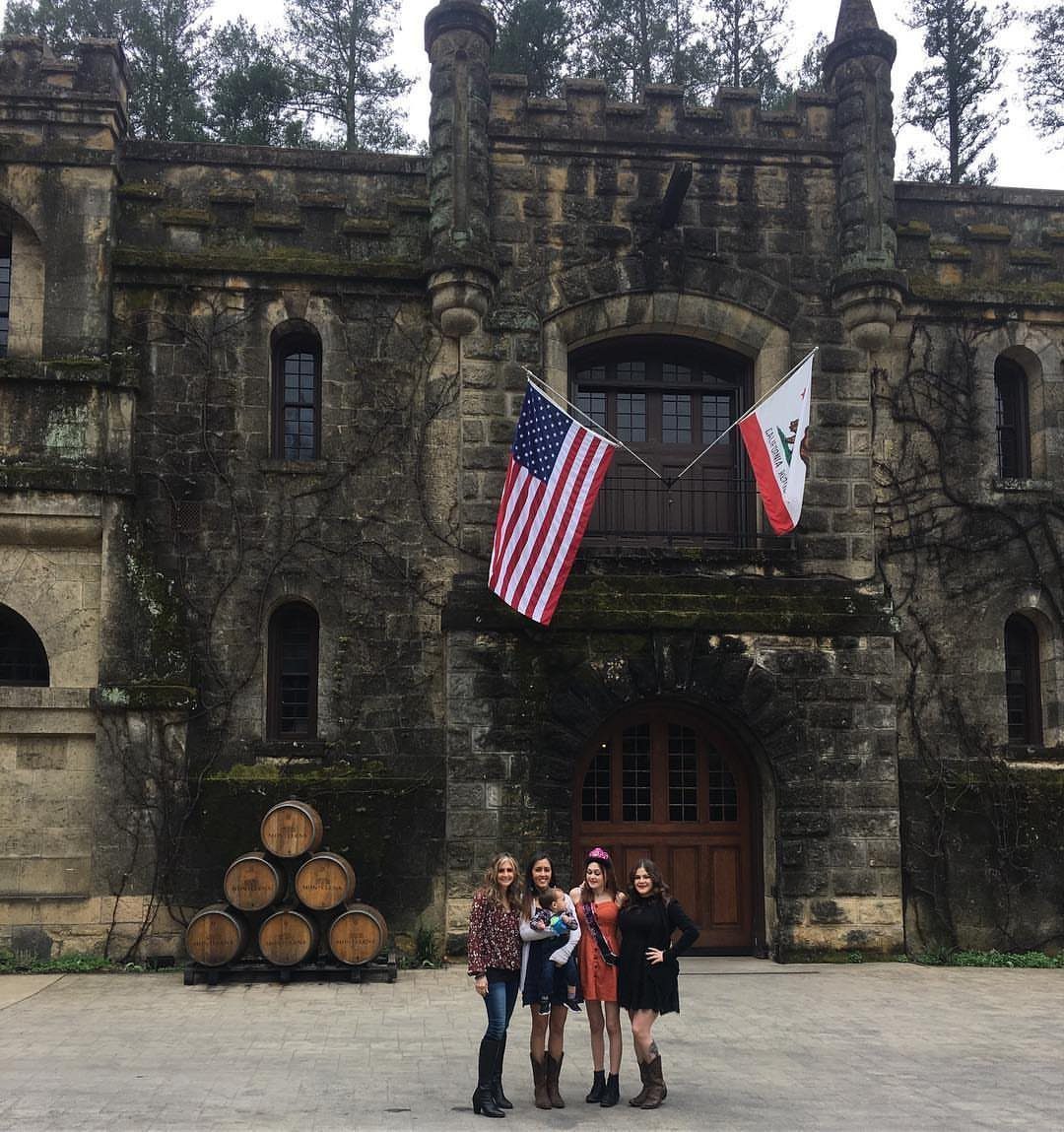 Four women posing in front of a stone castle with wooden wine barrels.