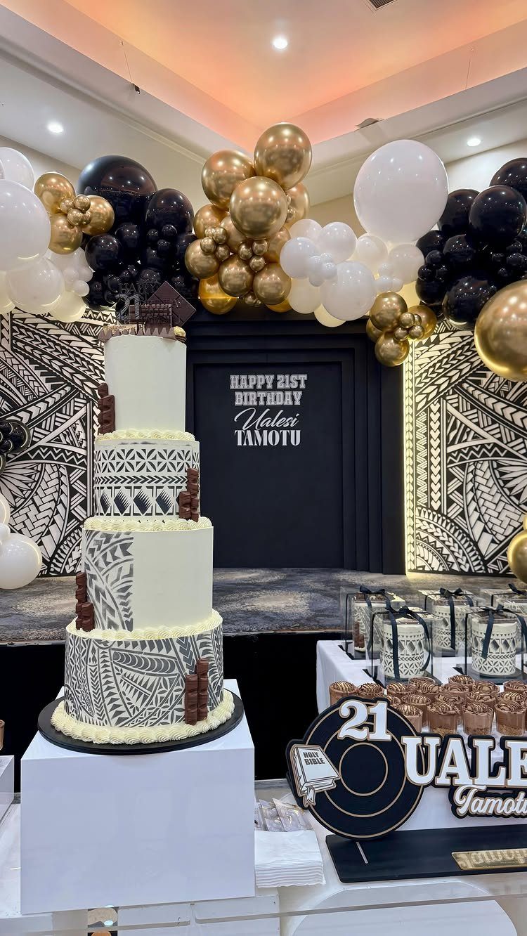 Four-tier white cake with tribal patterns and gold, black, and white balloons.