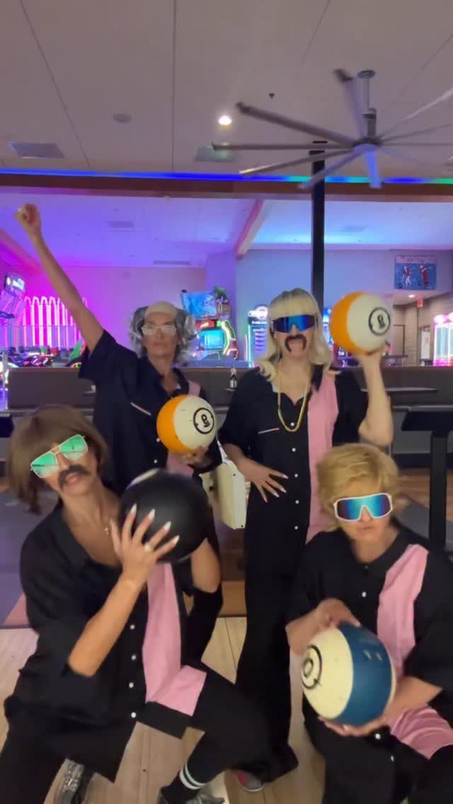 Four friends in matching bowling shirts and wigs posing with bowling balls.