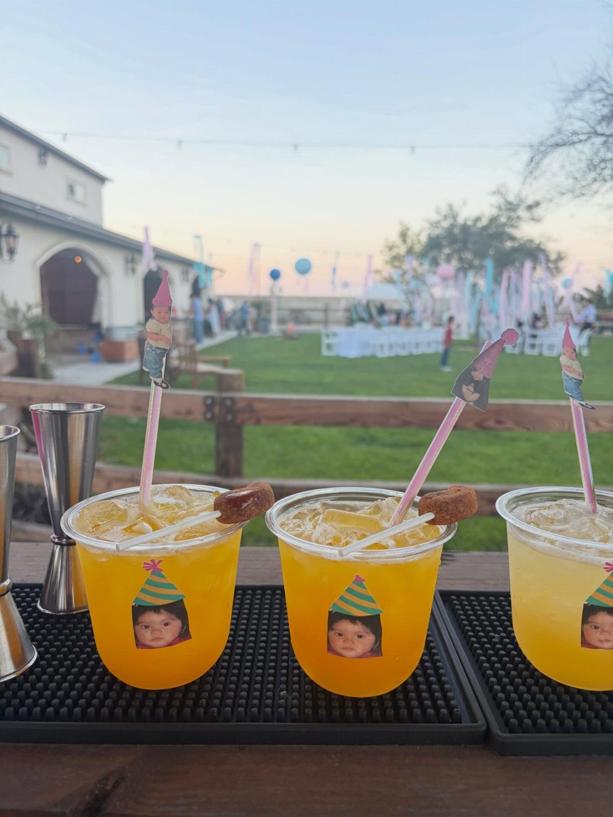 Yellow cocktails in plastic cups with baby photos and party hat stirrers outdoors