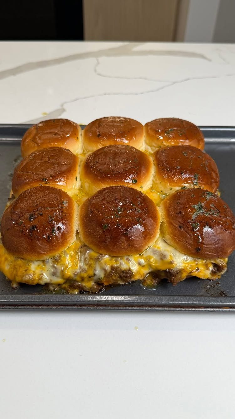 Cheesy meat sliders on a baking sheet atop a white marble surface.