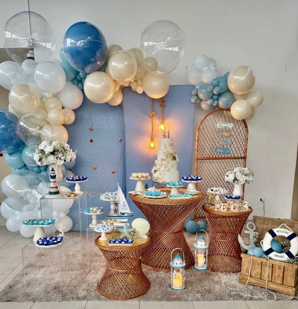 Nautical party decoration with blue balloons, wicker tables, and a white tiered cake.