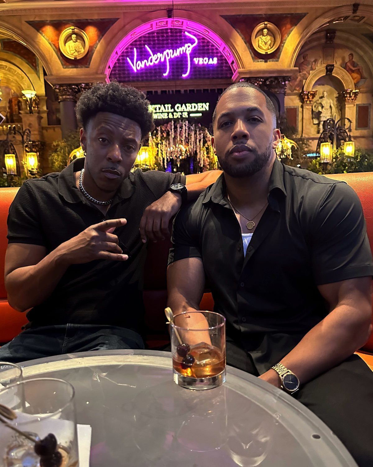 Two men in black shirts sitting at a lounge table with cocktails.