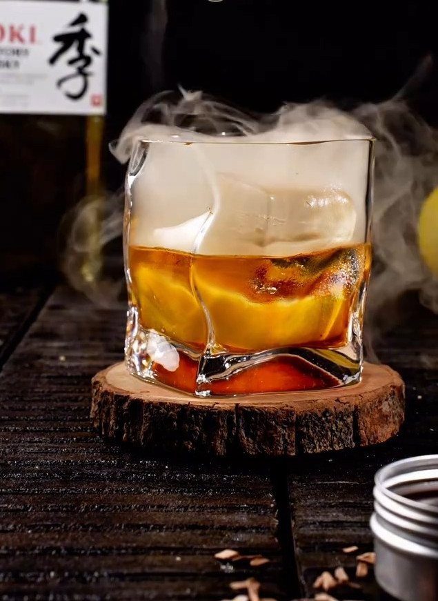 Amber whiskey with ice in a glass on a wooden slice coaster with smoke.
