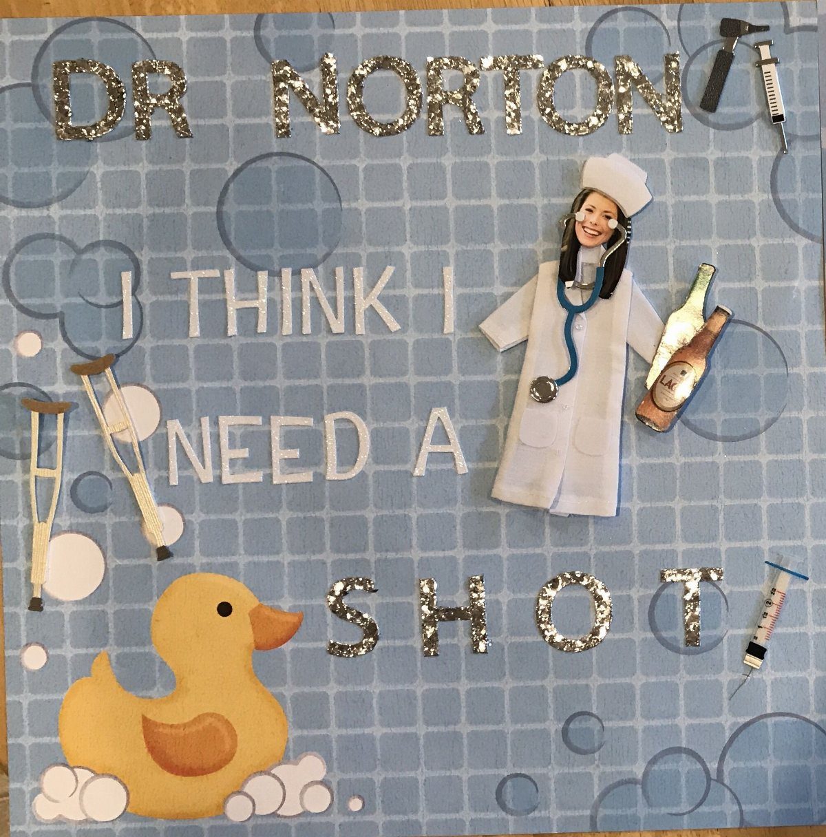Medical-themed scrapbook page with a paper doctor, glitter letters, and toy syringes.