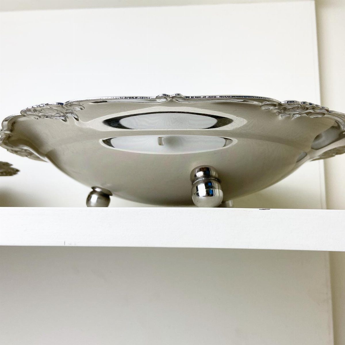 Silver decorative bowl with ornate edges resting on a white shelf