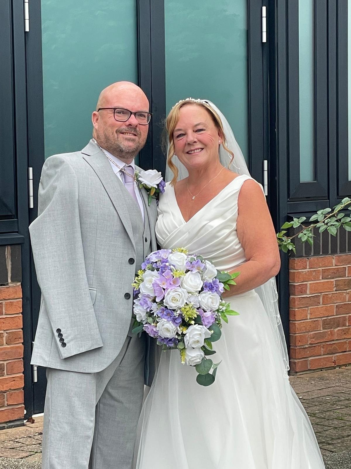 Smiling couple in wedding attire posing with a purple and white flower bouquet.