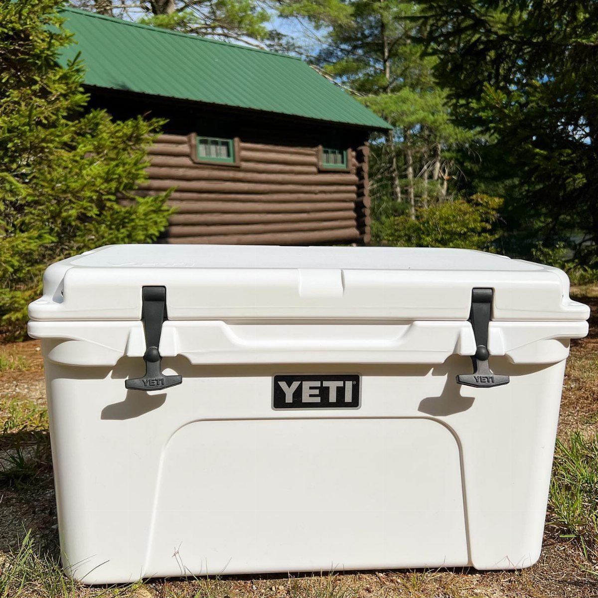 White Yeti cooler sitting on grass in front of a rustic log cabin.