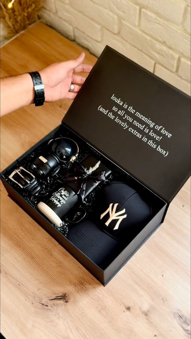 Black gift box containing a baseball cap, belt, watch, and mug on wood.