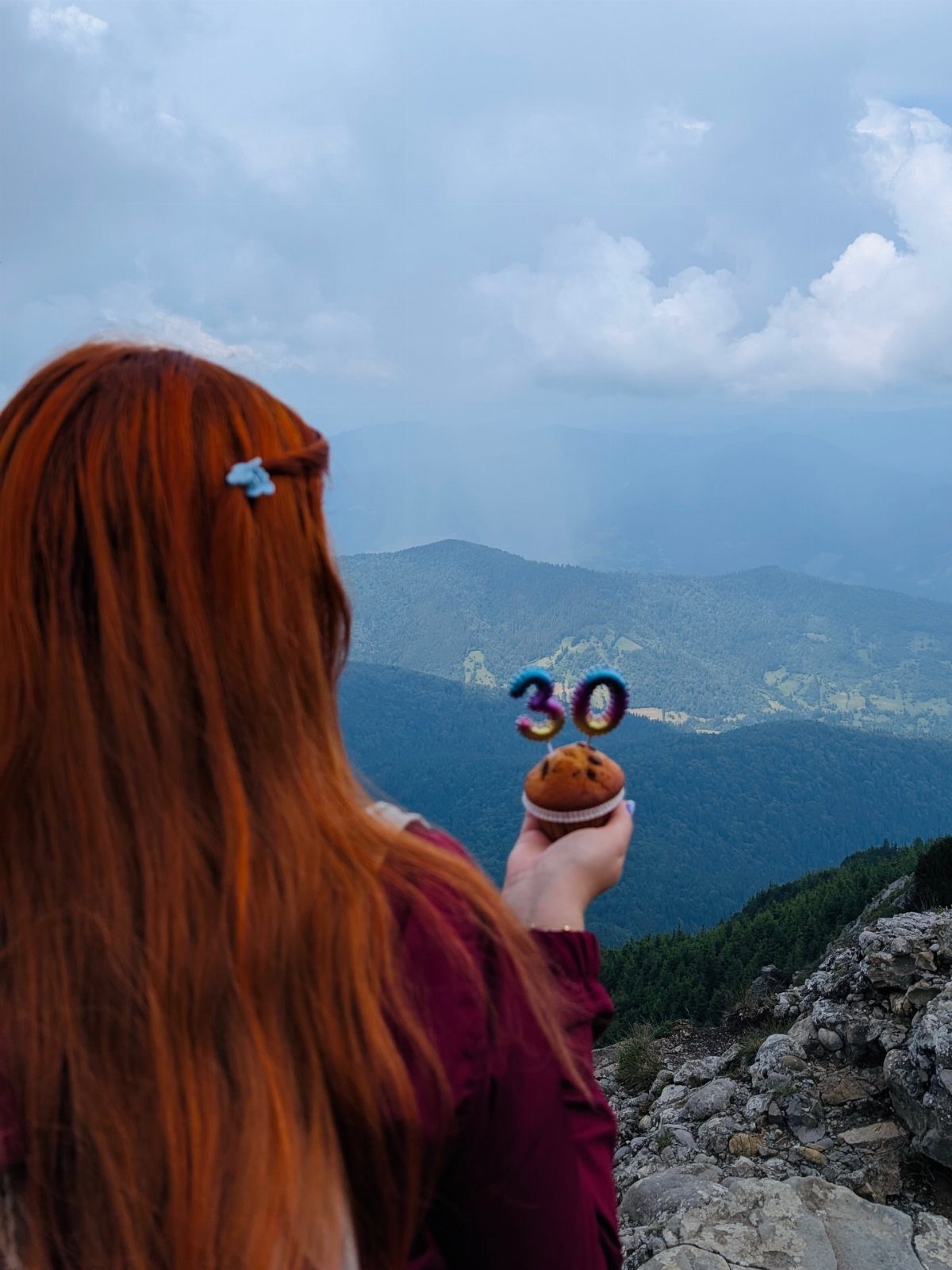 Woman with red hair holding a 30th birthday cake on a mountain peak