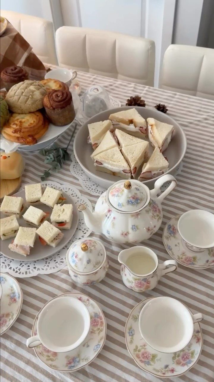 Floral teapot and teacups with finger sandwiches and pastries on a striped tablecloth.