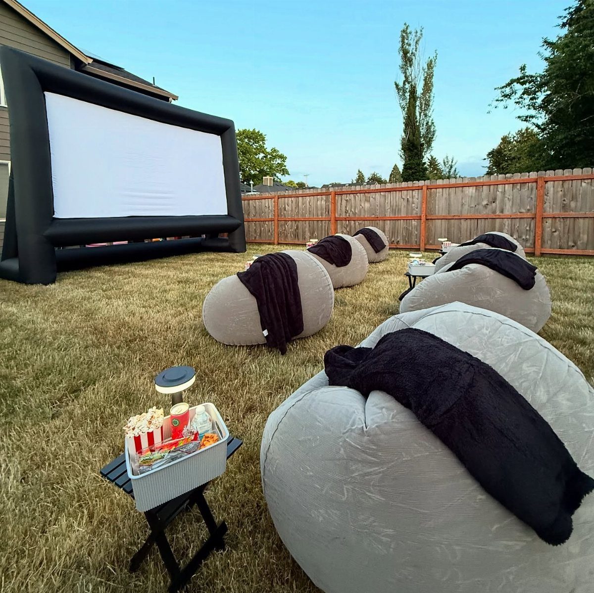 Outdoor movie screen and bean bag chairs with snacks in a grassy backyard.