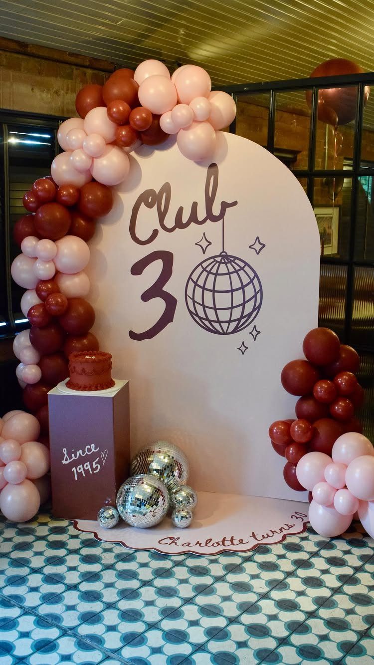Birthday party backdrop with Club 30 text, pink and burgundy balloons, and disco balls.