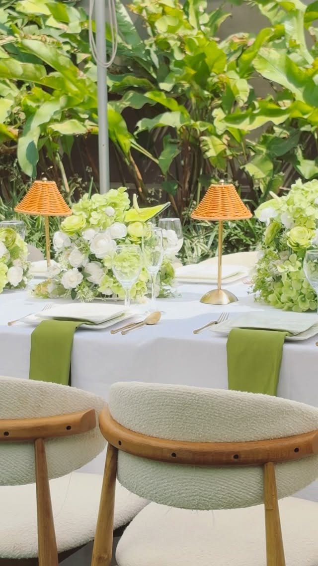 Outdoor dining table with white flowers and woven lamps set against tropical greenery
