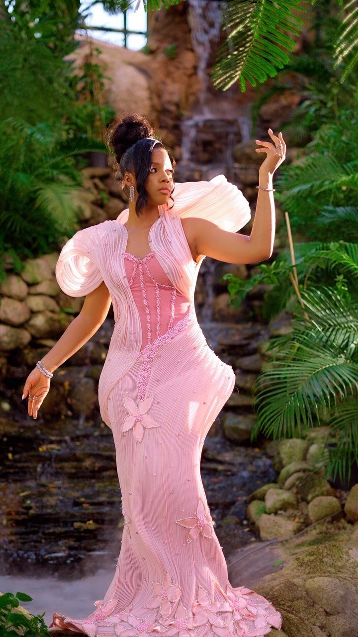 Woman in a detailed pink gown posing before a waterfall in a garden