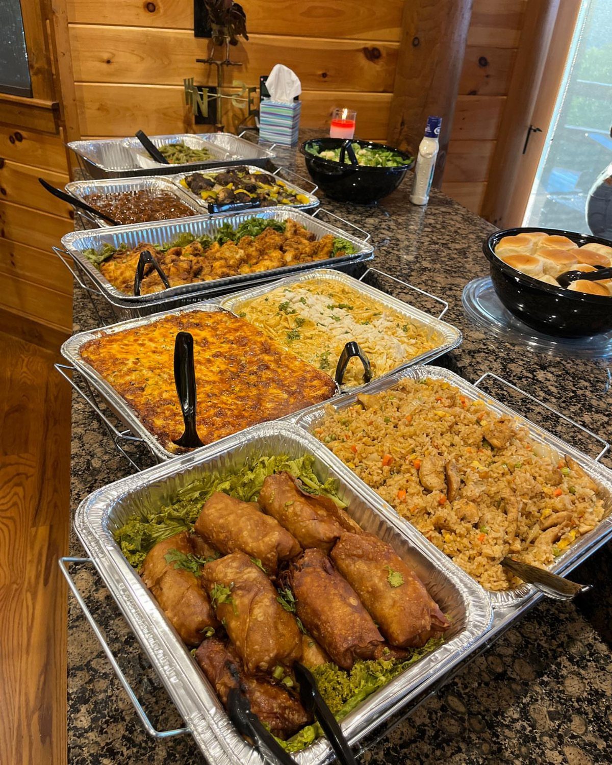 Assorted party foods in foil trays and bowls on a granite kitchen counter.