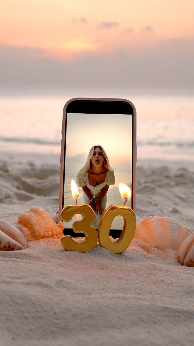 Gold number 30 candles lit in sand before a phone showing a woman.