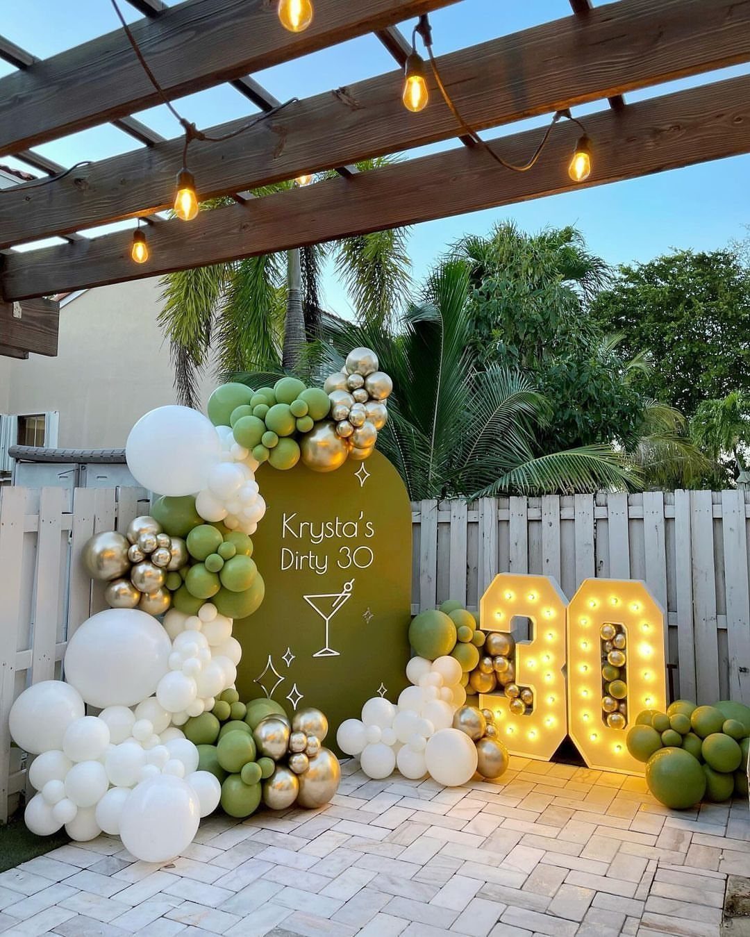 Party backdrop with green and gold balloons and glowing number 30 marquee lights.