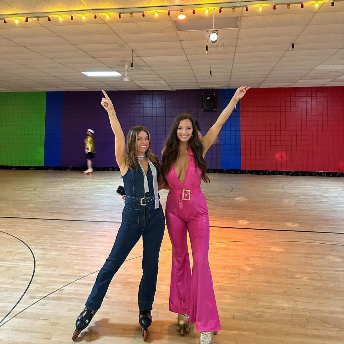 Two women in disco attire wearing roller skates posing on a wooden rink.