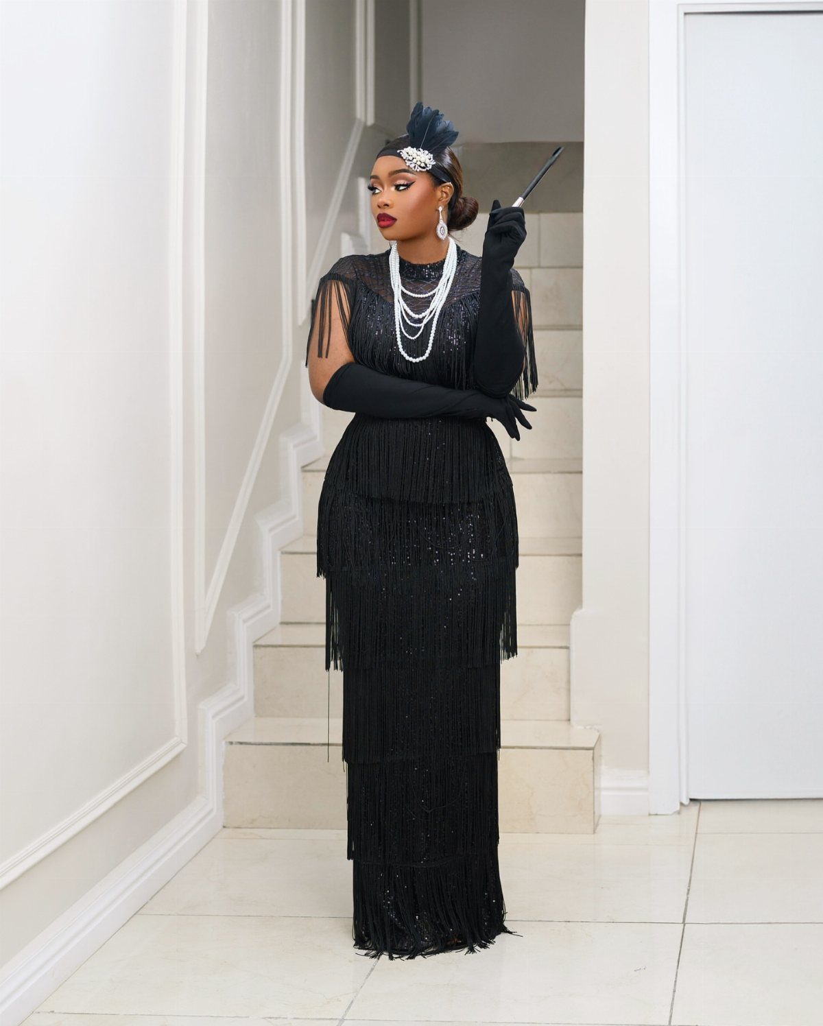 Woman in a black flapper dress with pearls and headband posing indoors.