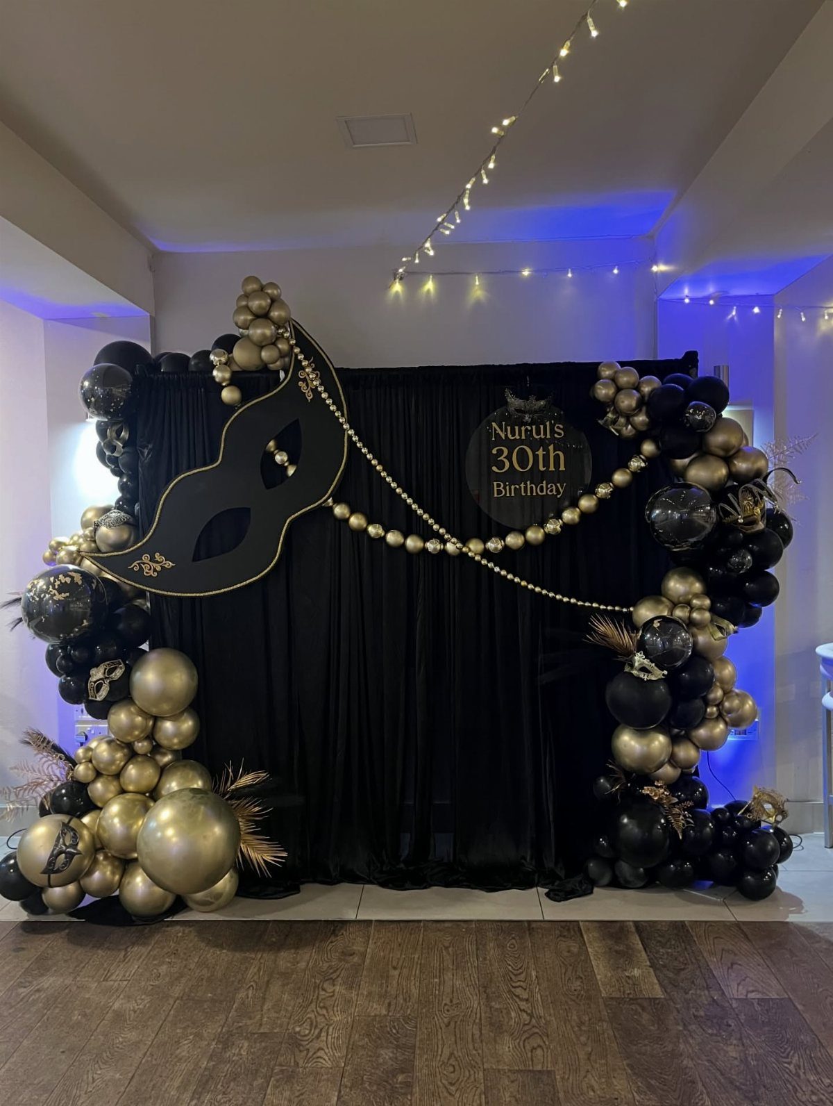 Black and gold birthday backdrop featuring a large masquerade mask and balloons.
