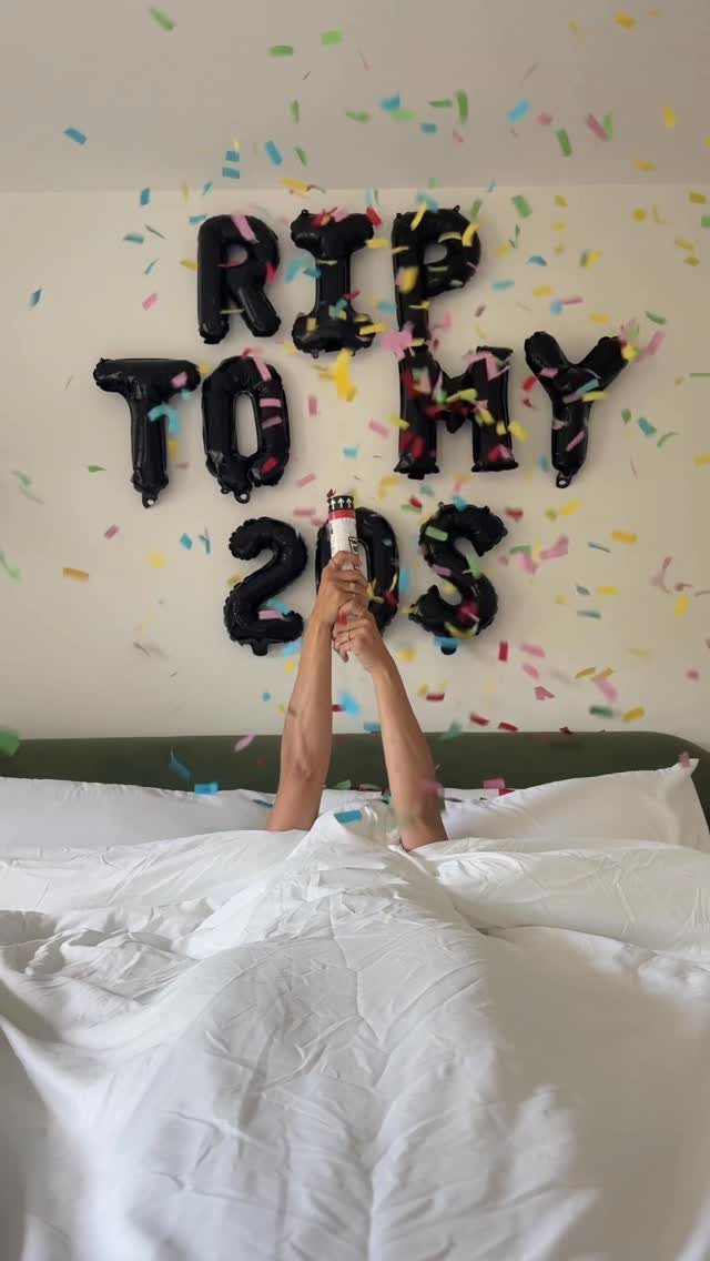 Person in bed with black balloons spelling RIP TO MY 20S and confetti