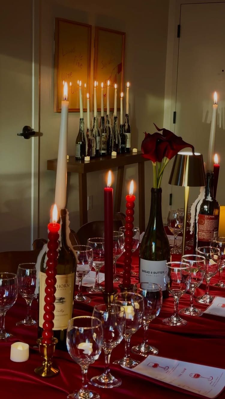 Elegant table setting with red tablecloth, wine glasses, bottles, and lit candles.