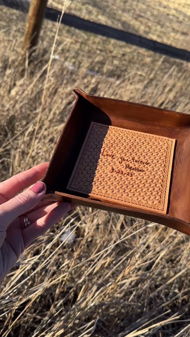 Brown leather catchall tray with personalized engraving held over dry outdoor grass.