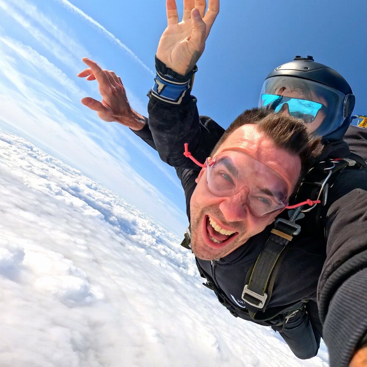 Man with glasses skydiving tandem above a sea of white clouds and blue sky.