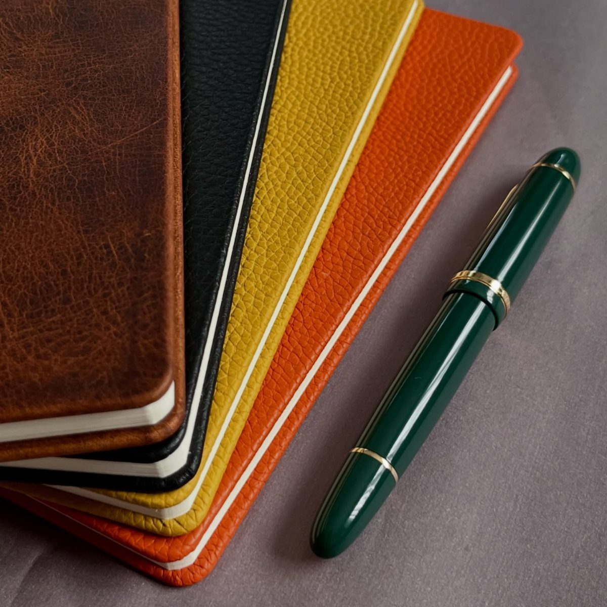 Leather notebooks in various colors with a green fountain pen on grey fabric