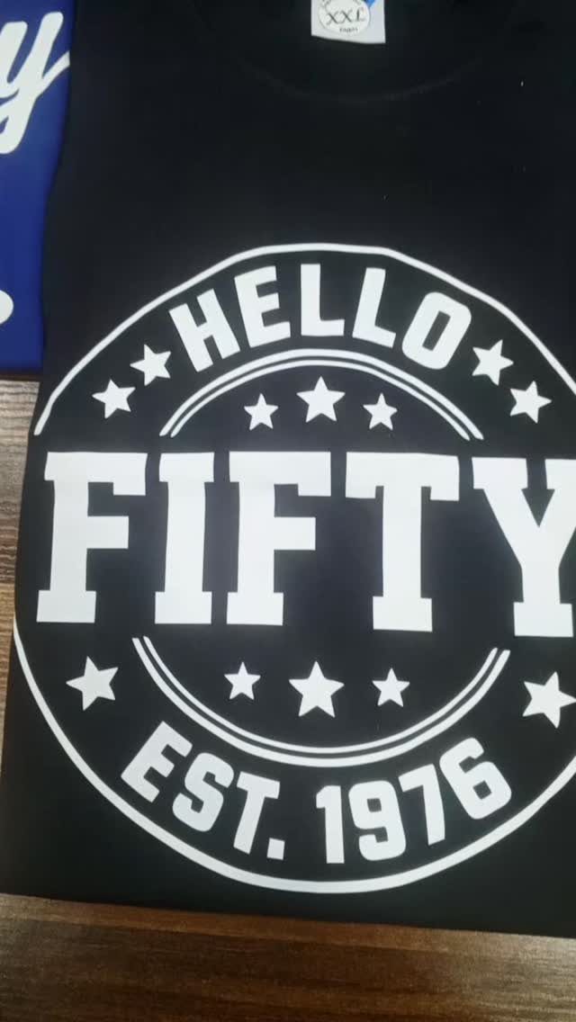 Black t-shirt with white text saying Hello Fifty Est. 1976 on wood.