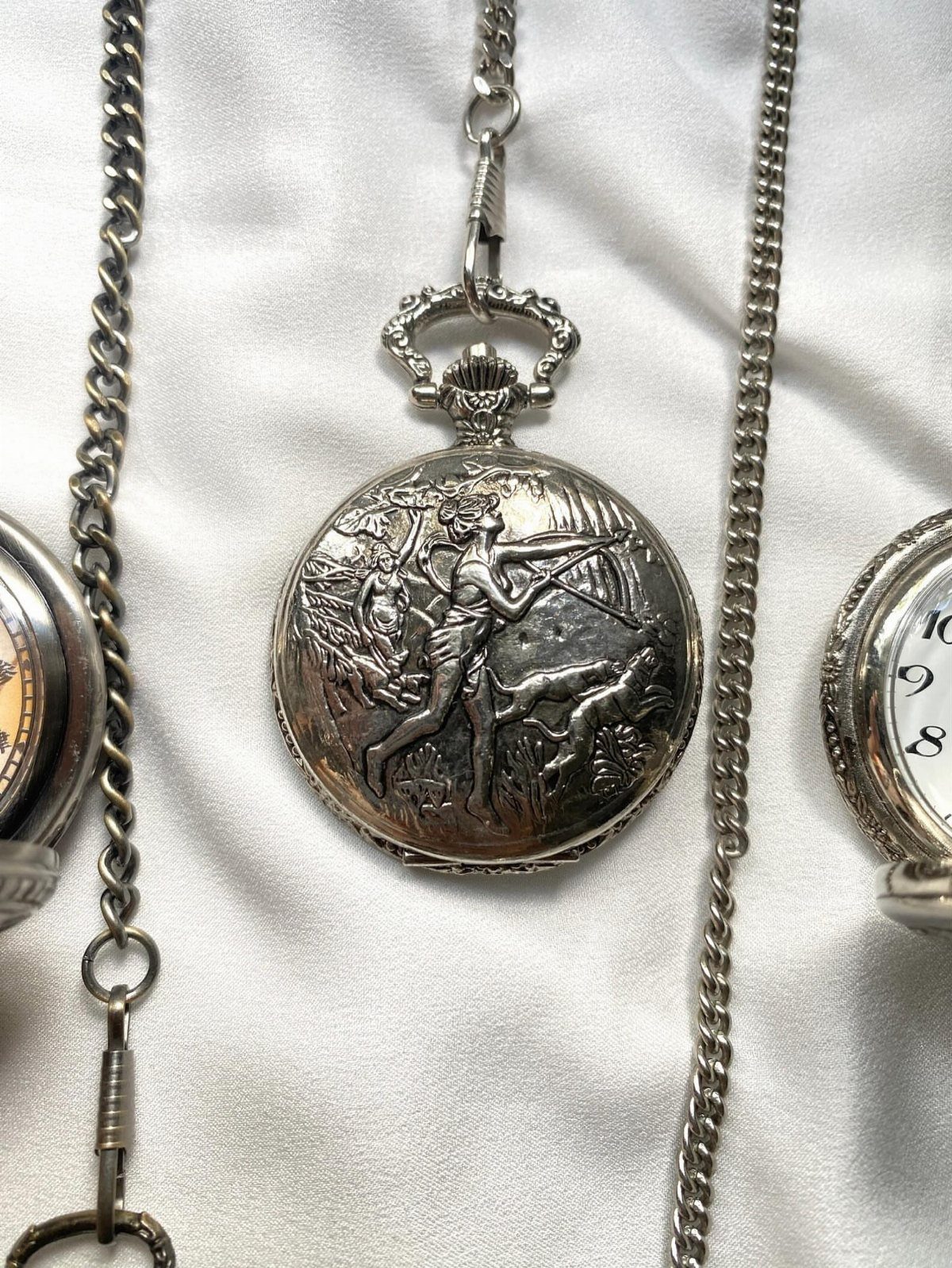 Silver pocket watches with ornate engravings and chains on white fabric