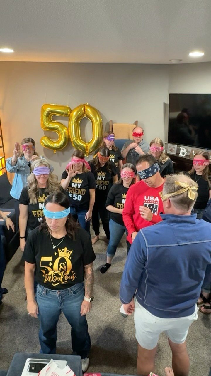 Group of blindfolded people playing a game near gold 50th birthday balloons.