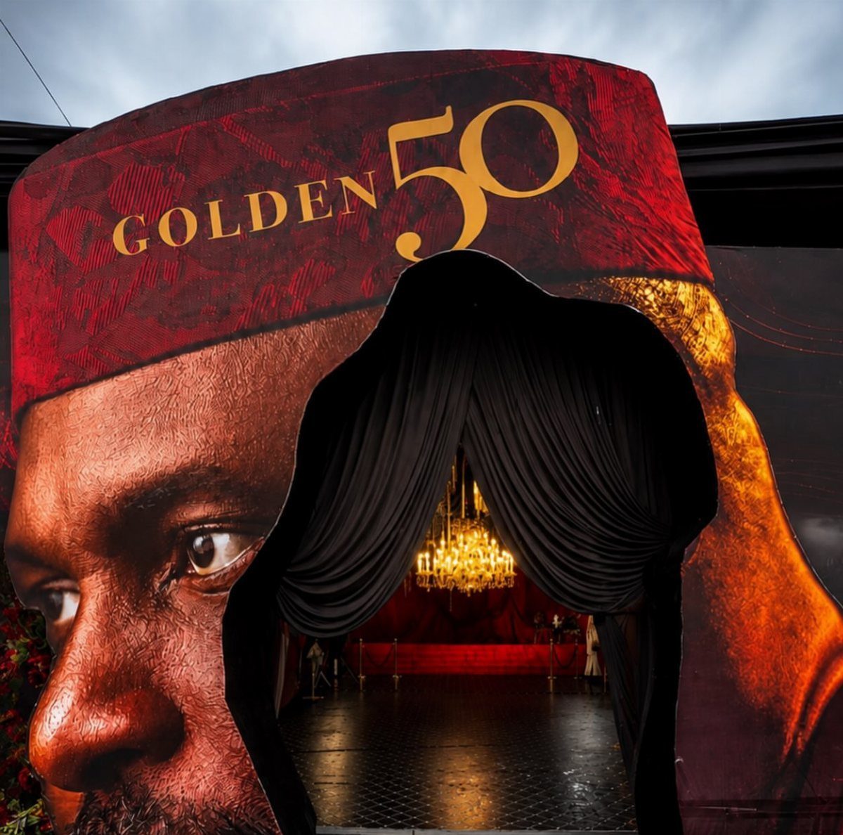 Event entrance with a large face cutout, gold text, and a crystal chandelier.
