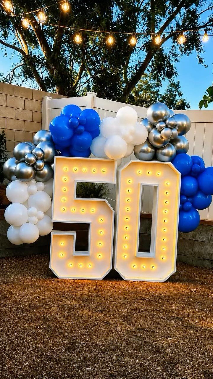 Large illuminated number 50 marquee signs surrounded by blue and silver balloons outdoors.